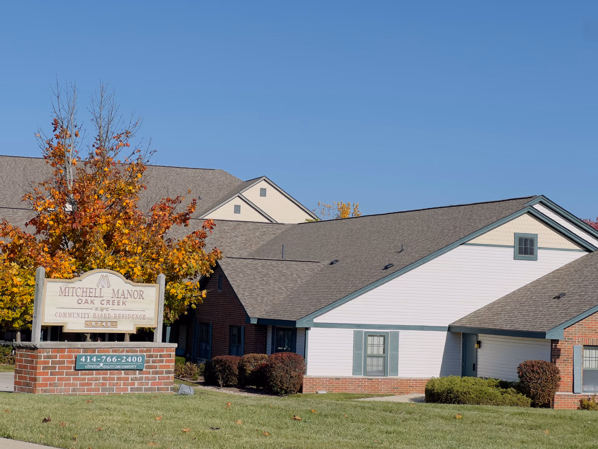 Exterior view of Mitchell Manor Oak Creek, a community-based residence. The building has a combination of brick and white siding with green trim and a gray shingled roof. In front of the building is a sign with the facility name, phone number, and a tree with autumn-colored leaves.