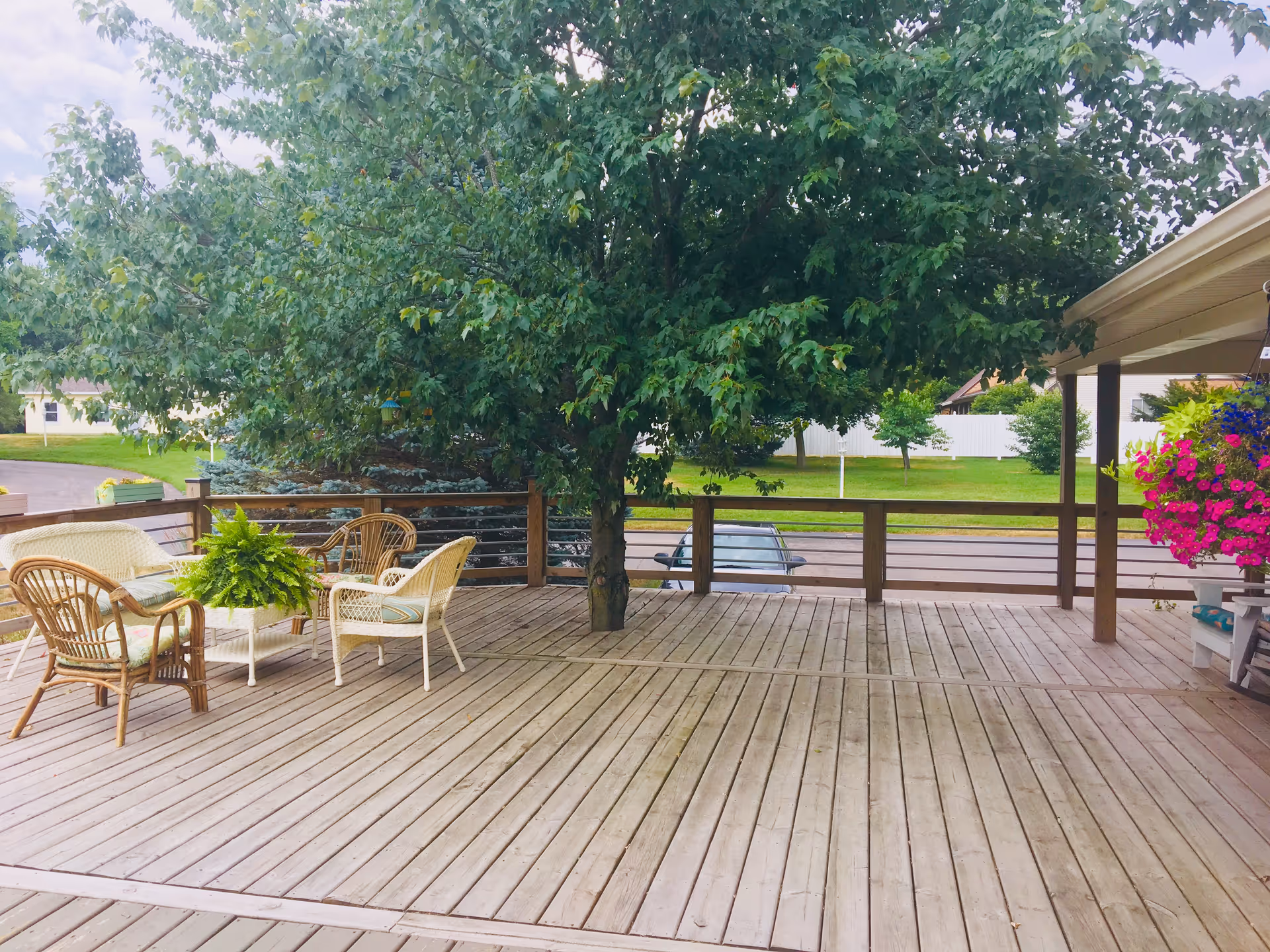 A spacious wooden outdoor deck with a large tree growing through the middle. The deck has a seating area with wicker chairs and a small table with a green potted plant. There is a hanging basket with vibrant pink and purple flowers on the right side under a covered section. In the background, there is a grassy area with trees and a white fence.