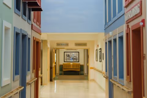 A brightly lit hallway in a healthcare facility with colorful walls on either side, handrails along the walls, and a bench at the end of the corridor beneath a framed picture of a bicycle. Signs above the bench indicate directions to 'New Residents' and 'Nursing Care'.