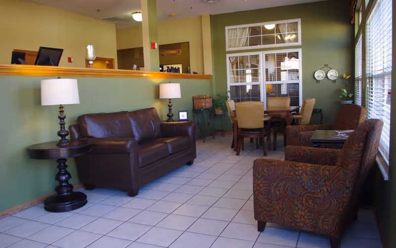 A bright lounge area with a leather sofa, patterned armchairs, side tables and lamps, and a dining table near windows.