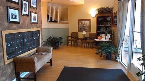 Interior view of a senior living facility lobby area with a row of mailboxes on the left wall, a bench below the mailboxes, a small seating area with two chairs and a table with a lamp in the background, plants, a bookshelf, and large glass doors on the right side leading outside.