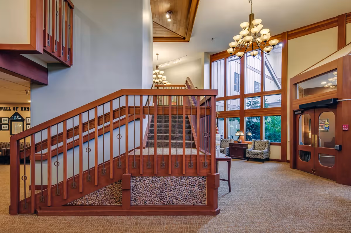 Interior view of a senior living facility lobby area featuring a carpeted staircase with wooden railings and stone accents underneath. The space has large floor-to-ceiling windows letting in natural light, two patterned armchairs with a side table and lamp, a chandelier hanging from the ceiling, and double wooden doors on the right side.