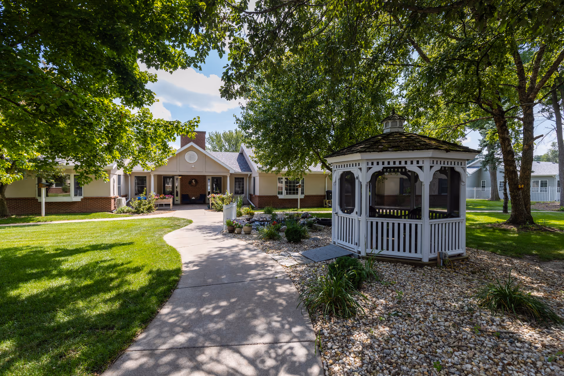 A sunny outdoor garden area at Country Manor Memory Care featuring a paved walkway leading to a single-story building with a brick chimney. On the right side of the walkway, there is a white wooden gazebo surrounded by rocks and plants. Large trees provide shade over the grassy lawn and walkway.