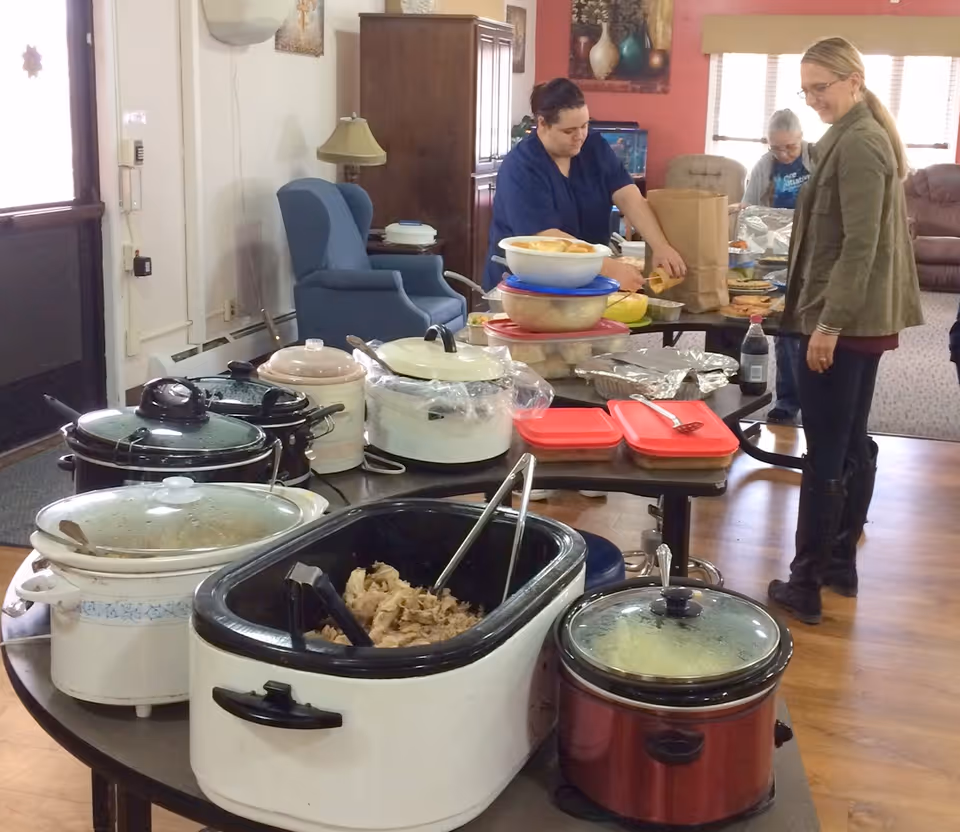 A room with several slow cookers and containers of food arranged on tables. Two women are standing near the tables, one wearing a blue uniform and the other in casual attire, preparing or serving food. The room has comfortable seating and a warm, home-like atmosphere.