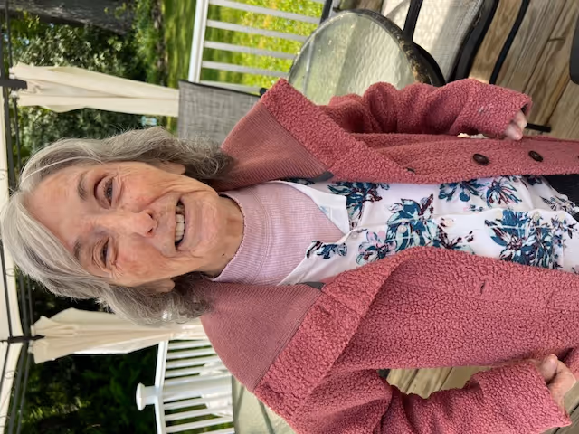 An elderly woman with gray hair wearing a pink textured coat over a floral patterned shirt, standing on a wooden deck with a glass table and chairs behind her, with greenery and trees in the background.