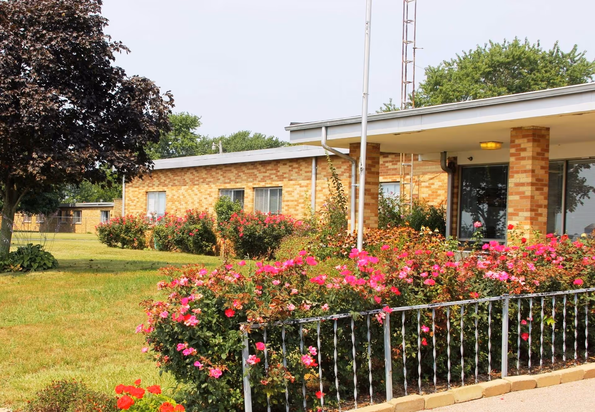 Exterior view of a single-story brick building with a covered entrance, surrounded by a garden with blooming pink and red flowers and a tree on the left side.