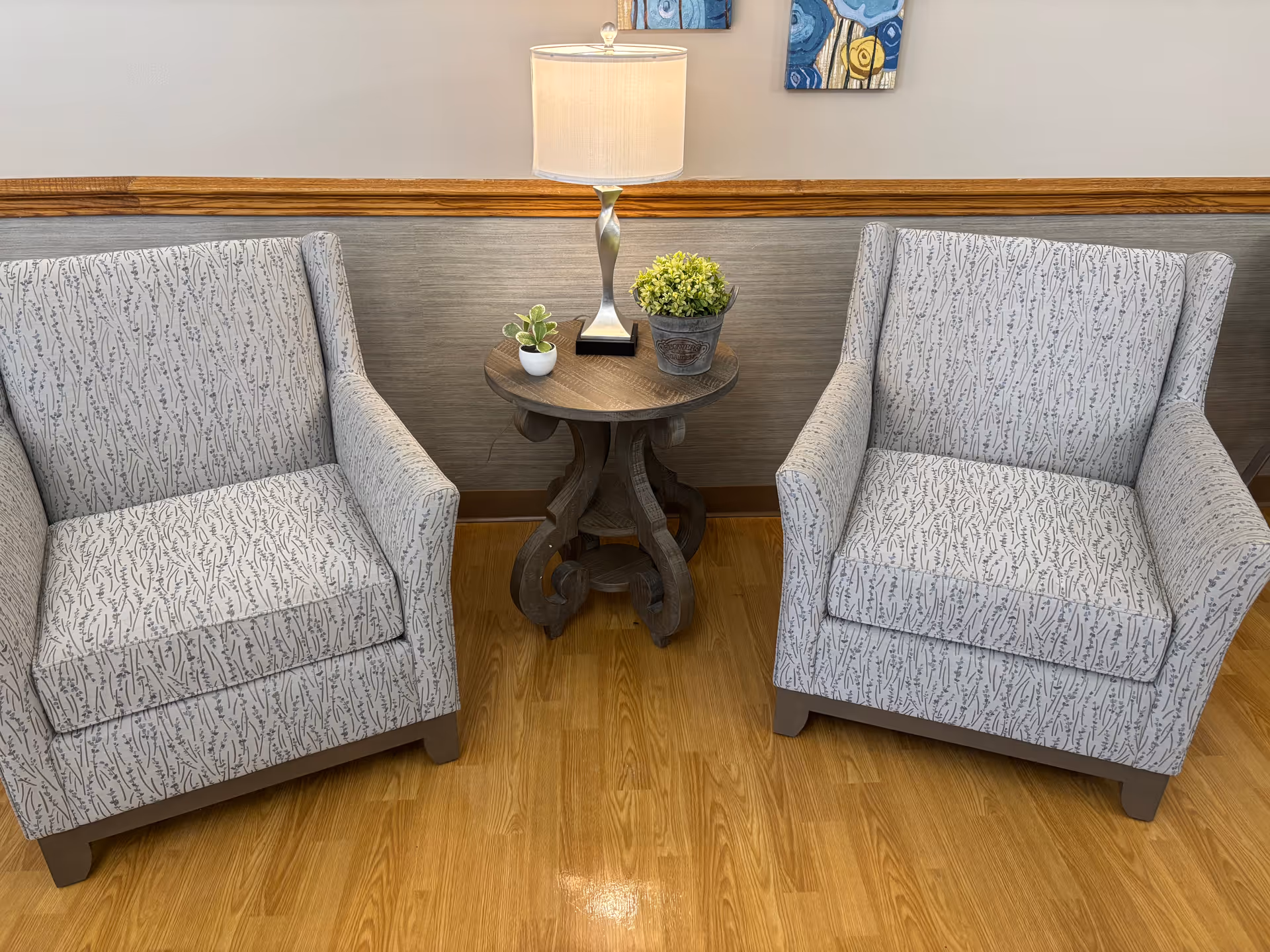 Two patterned armchairs with a small wooden side table between them. The table holds a lamp with a beige shade and two small potted plants. The setting is indoors with wooden flooring and a gray wall with wooden trim.