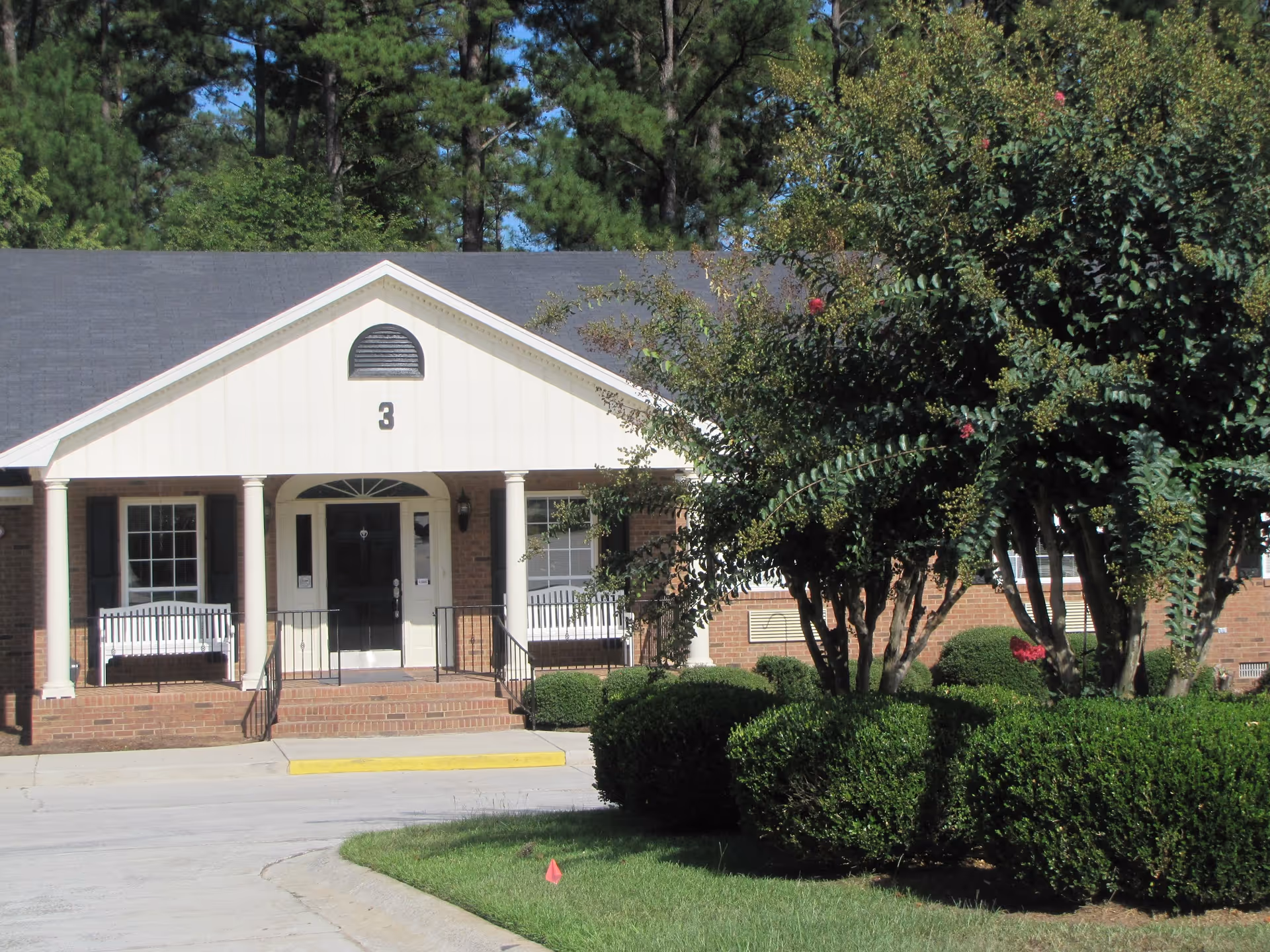 Front entrance of a low brick building with columns, a '3' above the doorway, porch benches and landscaped shrubs and trees.