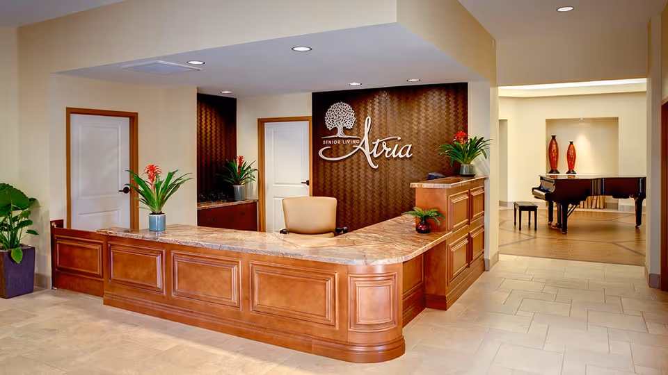 Reception area of Atria Hacienda senior living facility featuring a wooden front desk with a marble countertop, potted plants on the desk, a beige chair behind the desk, and a wall with the Atria Senior Living logo. In the background, there is a hallway leading to a room with a black grand piano and decorative vases.