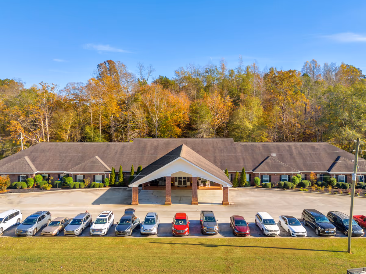 Front view of a single-story senior living building with a covered entrance, parked cars in front, and trees with autumn foliage behind.