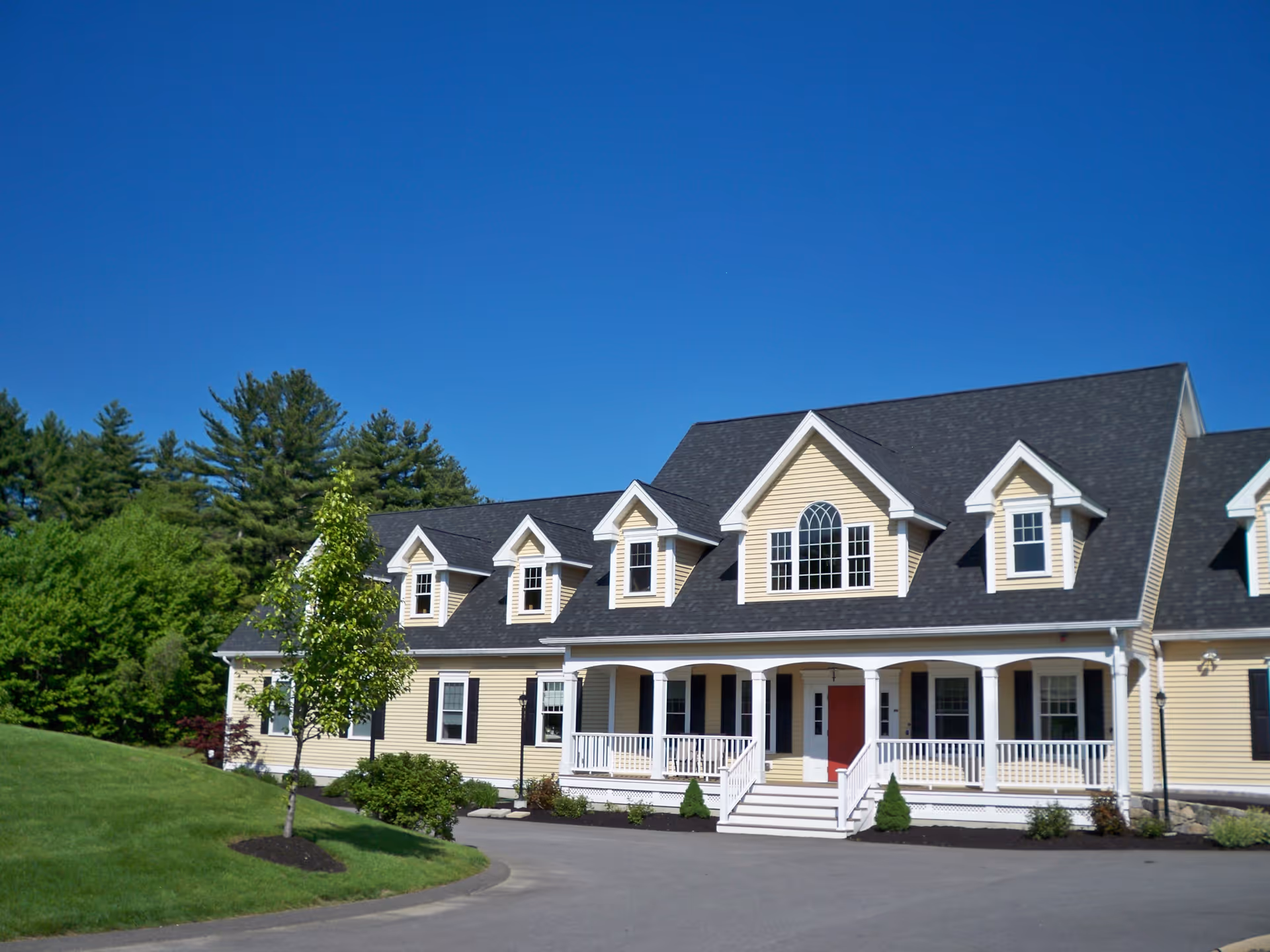A large, yellow two-story house with a dark gray roof and white trim. The house features multiple dormer windows and a covered front porch with white railings. The building is surrounded by a well-maintained lawn, small trees, and shrubs under a clear blue sky.