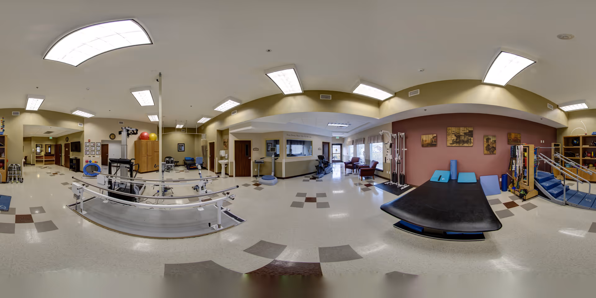 A spacious physical therapy and rehabilitation room in Sunnyside Healthcare Center featuring parallel bars for walking exercises, therapy mats, exercise balls, various rehabilitation equipment, and seating areas. The room is well-lit with overhead fluorescent lights and has a clean, organized appearance with beige and maroon walls.