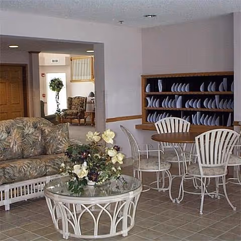 Communal lounge area with wicker sofas, a glass-top coffee table with a floral centerpiece, and a dining table with chairs beside a wall rack of plates.