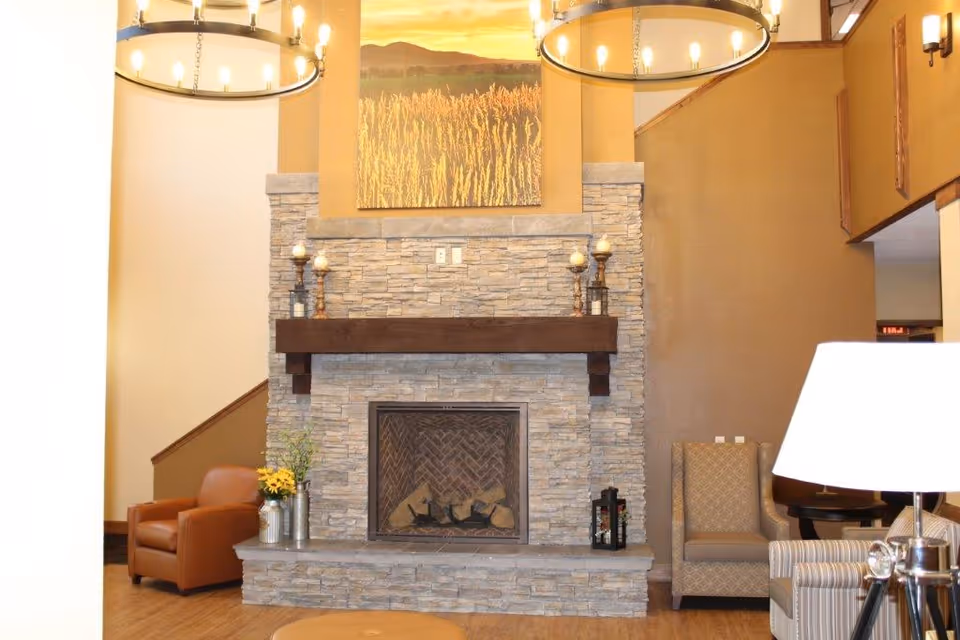 A cozy living room area featuring a stone fireplace with a wooden mantle, decorated with candles and a lantern. Above the fireplace is a large painting of a field with mountains in the background. The room includes comfortable seating such as a brown leather armchair, a patterned armchair, and a striped armchair. Two large circular chandeliers hang from the ceiling, and a floor lamp is visible on the right side.