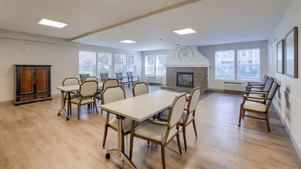 A spacious, well-lit common area with multiple tables and chairs arranged for seating. The room features large windows allowing natural light, a fireplace with a clock above it, and additional chairs lined up against the walls. The flooring is light wood, and the walls are painted in neutral tones.