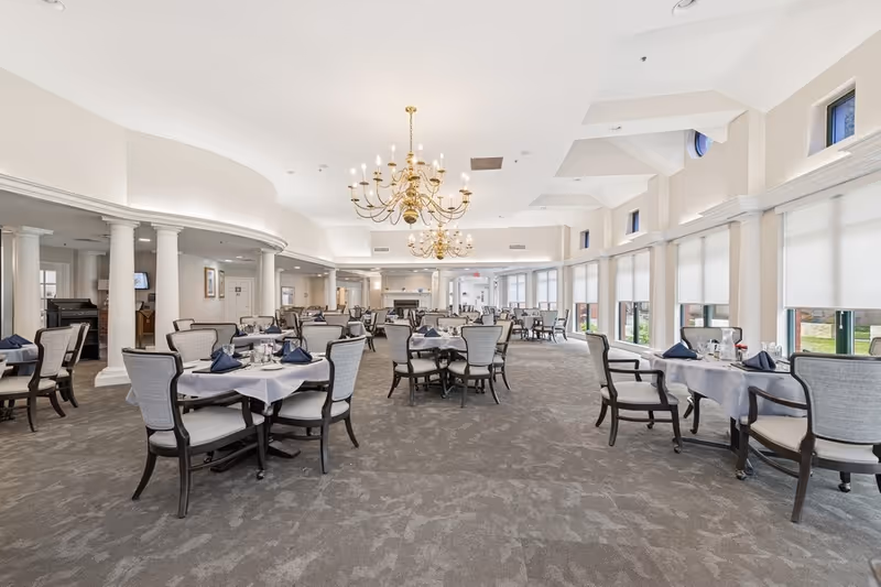 Spacious dining room with multiple round tables covered with white tablecloths and set with navy blue napkins and glassware. The room features large windows with white blinds, beige walls, carpeted floor, white columns, and elegant gold chandeliers hanging from the ceiling.