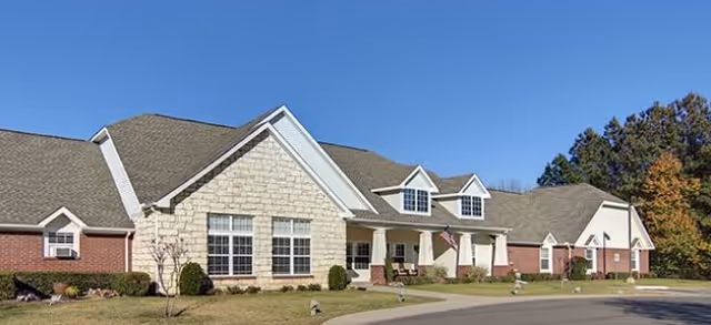 Single-story brick and stone senior living building with a covered entrance, American flag, driveway and lawn under a clear blue sky.