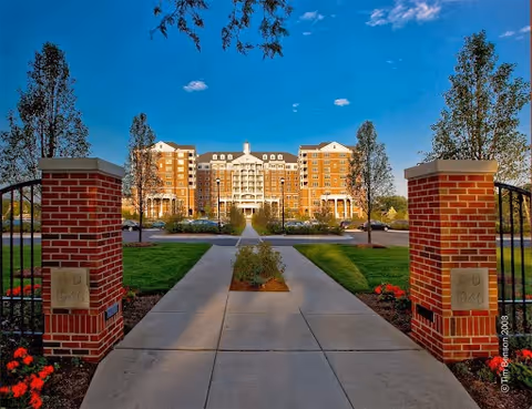 Wide concrete pathway leading through a landscaped garden area with red flowers and young trees, flanked by two brick pillars, toward a large multi-story residential building under a clear blue sky.