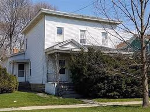 White two-story building with a small front porch, stairs, and trees in the yard.