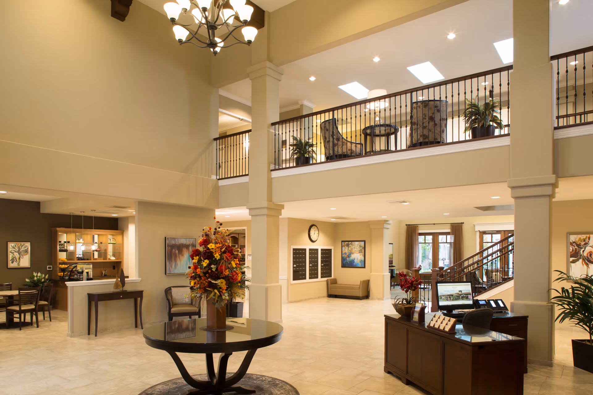 Spacious and well-lit lobby area of a senior living facility with a high ceiling, a round table with a large floral arrangement in the center, a reception desk with a computer, seating areas, wall art, and a staircase leading to an upper level with additional seating and plants.