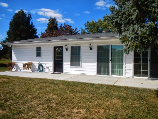 Single-story white building exterior with a black entry door, sliding glass doors, a concrete patio with chairs and a lawn in front.