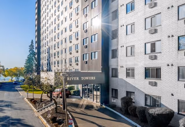 Exterior view of a multi-story residential building named River Towers with a canopy over the entrance. The building is white with many windows and air conditioning units. There are trees, shrubs, and a paved driveway leading to the entrance. The sun is shining brightly, reflecting off the building.