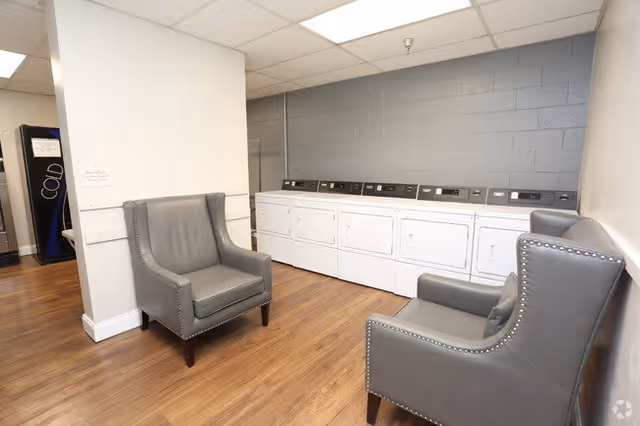 A laundry room with a row of white washing machines and dryers against a gray brick wall. Two gray upholstered armchairs with nailhead trim are placed on a wooden floor, facing each other. A vending machine is visible in the background.