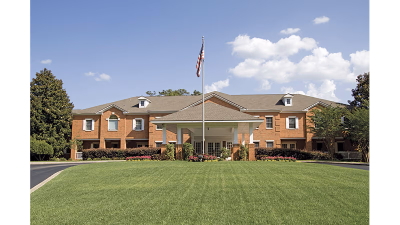 Front exterior of a two-story brick senior living building with a covered entrance, flagpole, and expansive lawn.