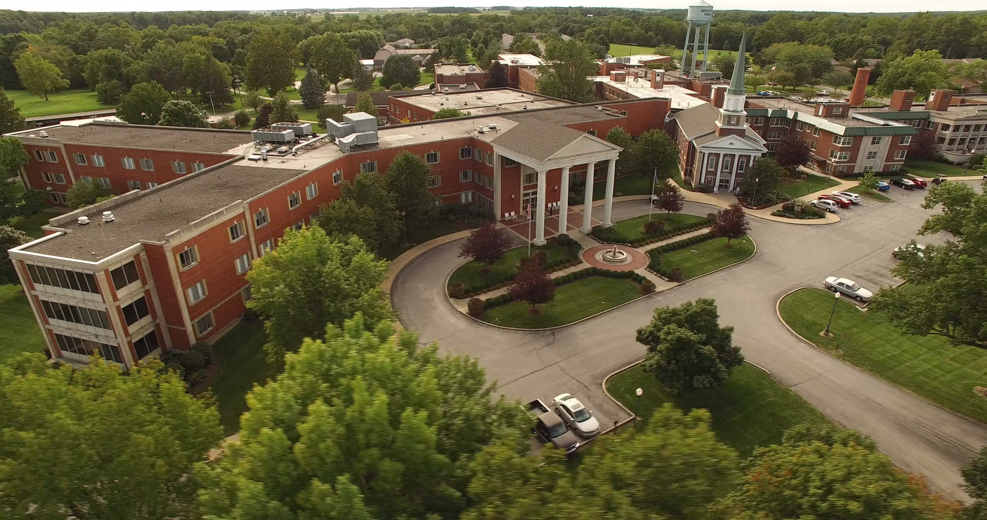 Aerial view of Heritage Pointe of Warren, a large senior living facility with multiple connected brick buildings, a circular driveway with a fountain, surrounded by green trees and lawns, and a water tower in the background.