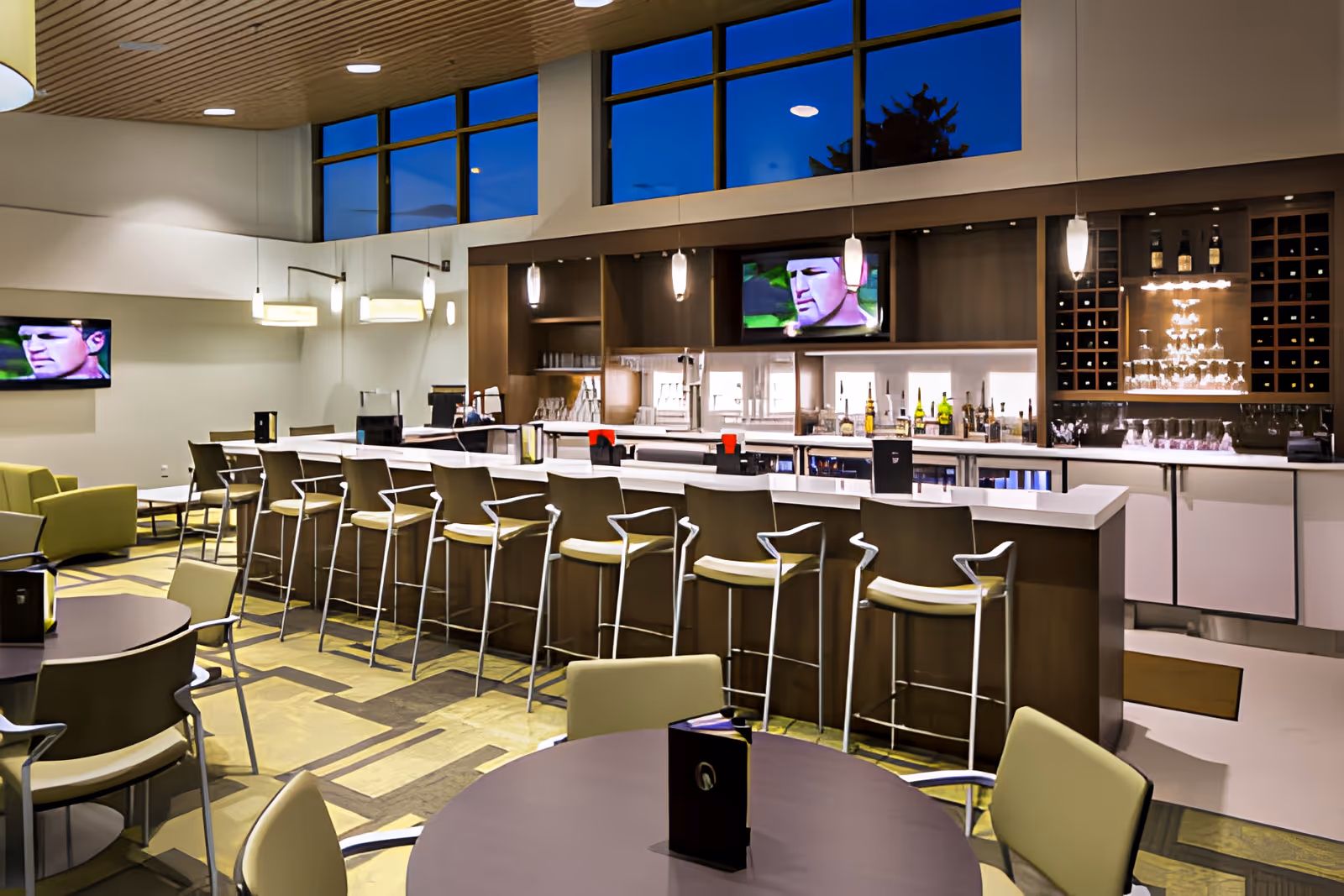 Modern communal dining and bar area with high stools, tables, lounge seating, and wall-mounted TVs beneath large windows.
