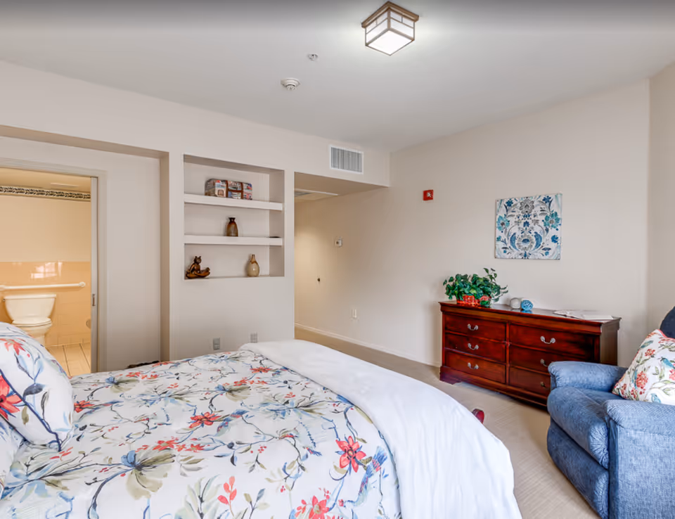 A bright bedroom with a floral patterned bedspread on the bed, a blue armchair with a floral pillow, a wooden dresser with decorative items and a plant on top, and a wall painting above the dresser. There is an open doorway leading to a bathroom with a visible toilet and grab bars. The room has light-colored walls and carpeted floor.