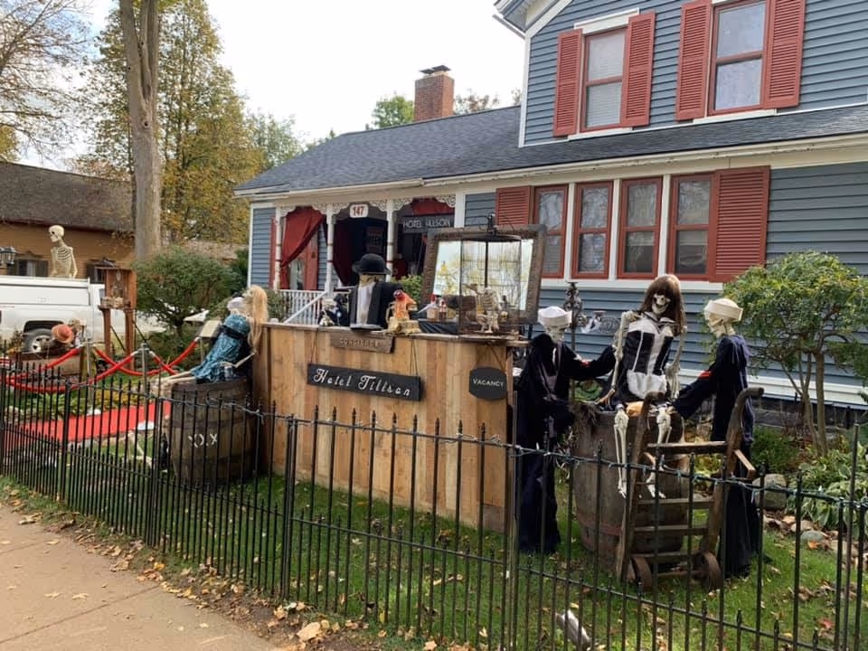 Front yard of a gray house with red shutters decorated with a Halloween skeleton display and a wooden 'Hotel Tillson' sign behind a black iron fence.