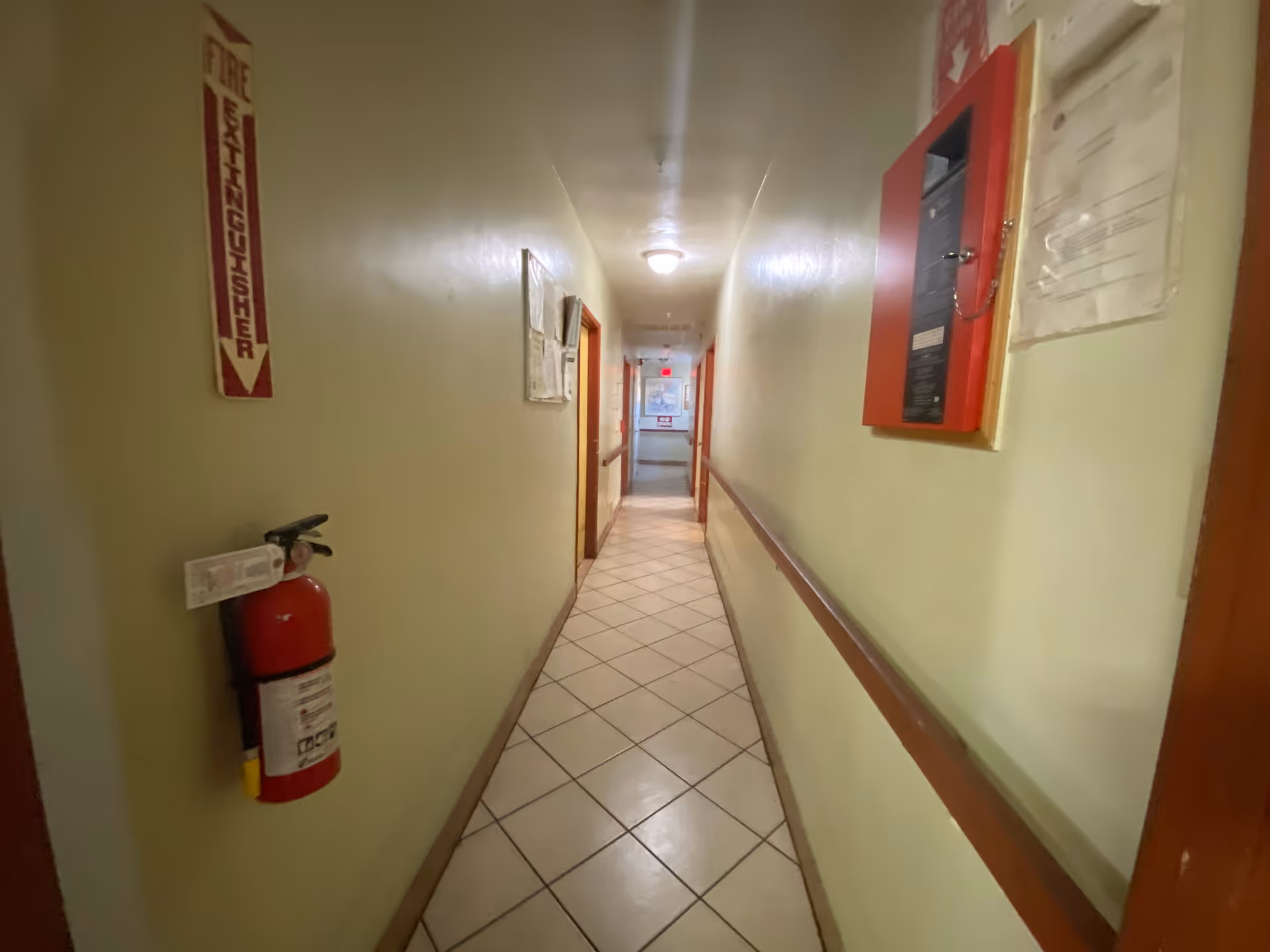 A narrow hallway with tiled floor and beige walls, featuring a fire extinguisher mounted on the left wall and a red fire alarm panel on the right wall. Several doors line the hallway, and a light fixture is on the ceiling.