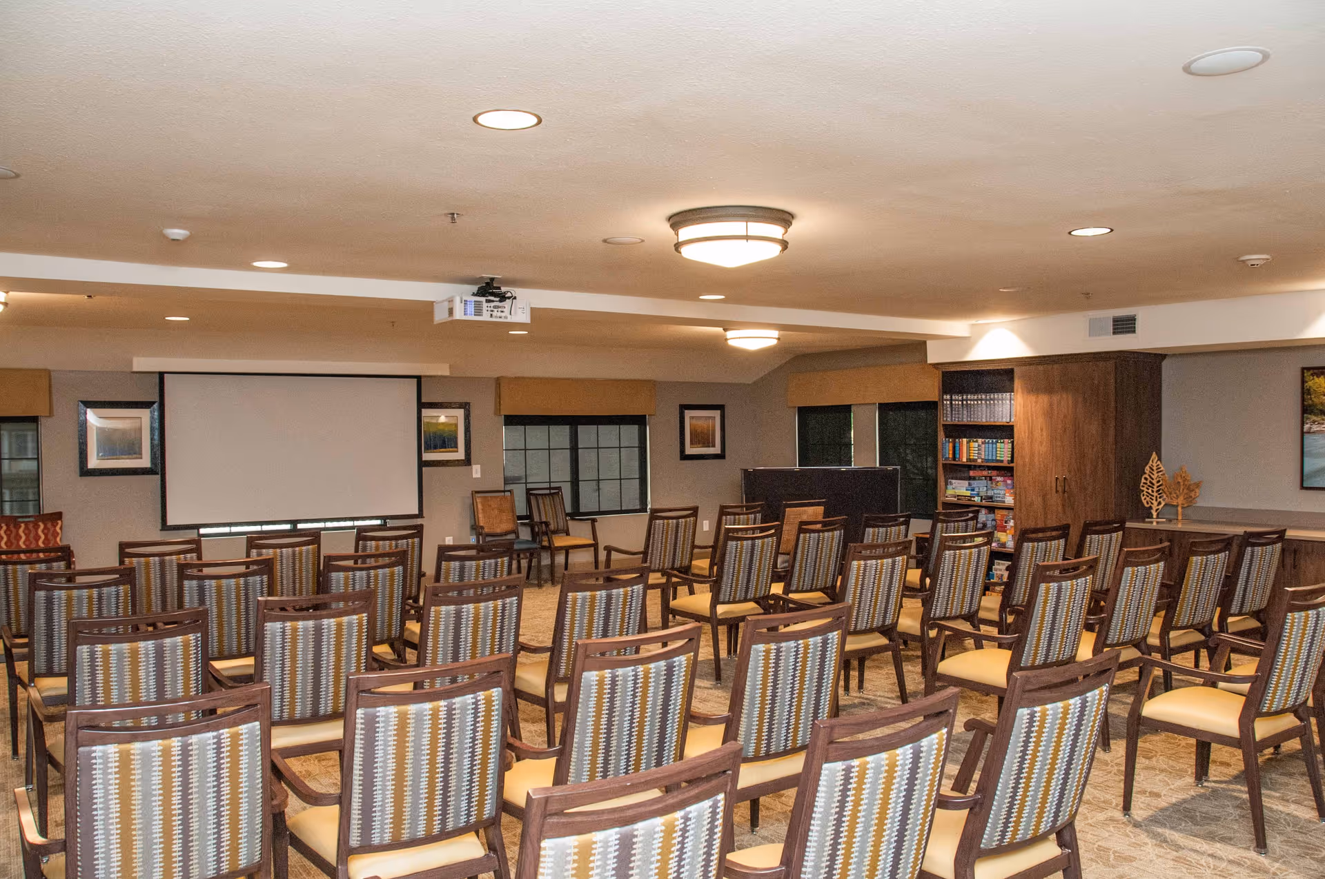 A spacious meeting or presentation room with rows of wooden chairs featuring patterned backs and cushioned seats. At the front of the room is a large projector screen mounted on the wall, with a ceiling-mounted projector above. The room has several windows with beige valances, framed artwork on the walls, and a bookshelf filled with books and games. The ceiling has recessed lighting and two circular light fixtures.