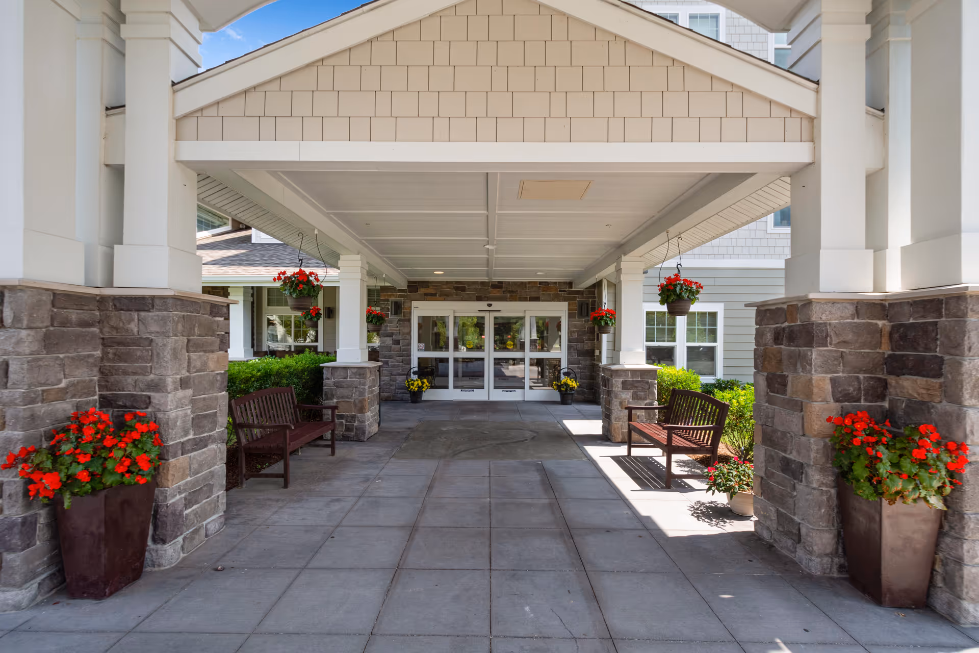Covered entrance to a senior living facility with stone pillars, hanging flower baskets with red flowers, two wooden benches on either side, and automatic glass doors at the end.