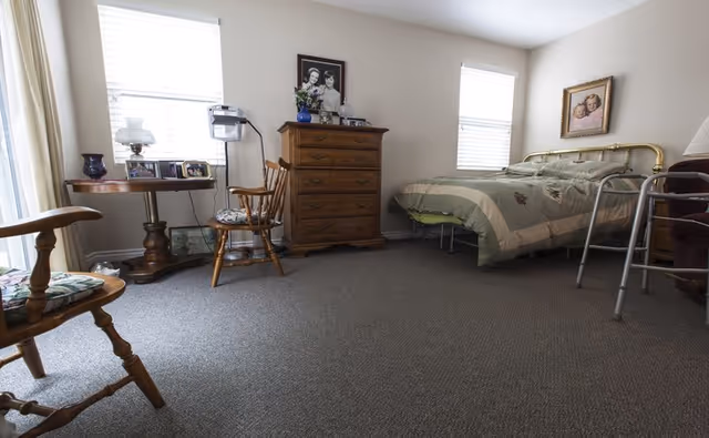 A bedroom with a brass bed frame, a green and beige quilt, a wooden dresser with framed photos and a vase with flowers, two wooden chairs with cushions, a small round wooden table with framed photos, and two windows with white blinds letting in natural light. A walker is positioned near the bed.