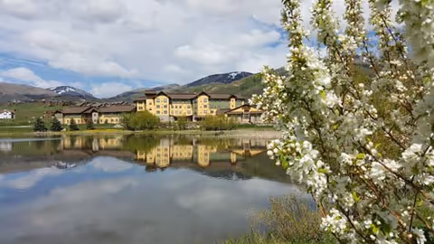 A large senior living facility named Casey's Pond is seen across a calm pond reflecting the building and surrounding landscape. In the foreground, there are blooming white flowers on a tree branch. The background features mountains under a partly cloudy sky.