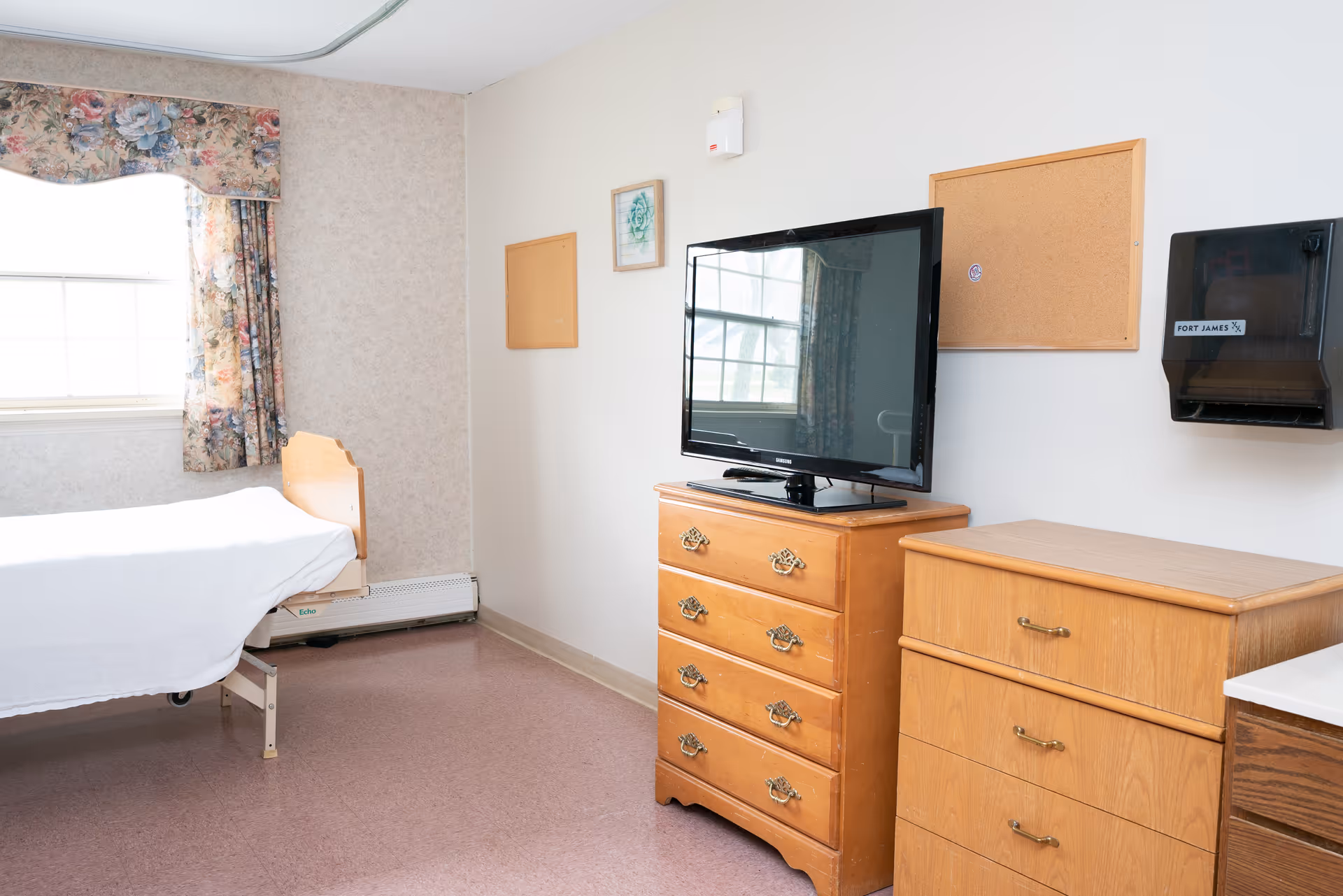 A simple room with a single bed covered with a white sheet, a wooden dresser with a flat-screen TV on top, another wooden chest of drawers, a window with floral curtains, and two empty cork boards on the wall.