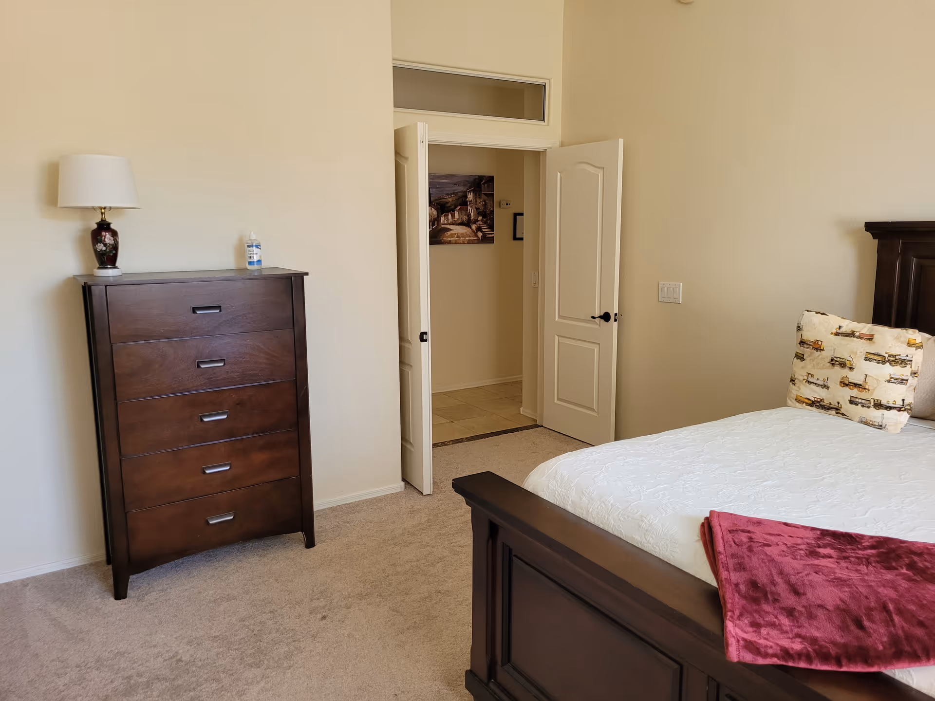 A bedroom with a dark wooden bed frame and a white bedspread. There is a pillow with a truck pattern and a folded red blanket on the bed. To the left, there is a tall dark wooden dresser with a lamp and a bottle on top. The room has beige walls and carpet, and a door is open leading to another room with tiled flooring and a picture on the wall.