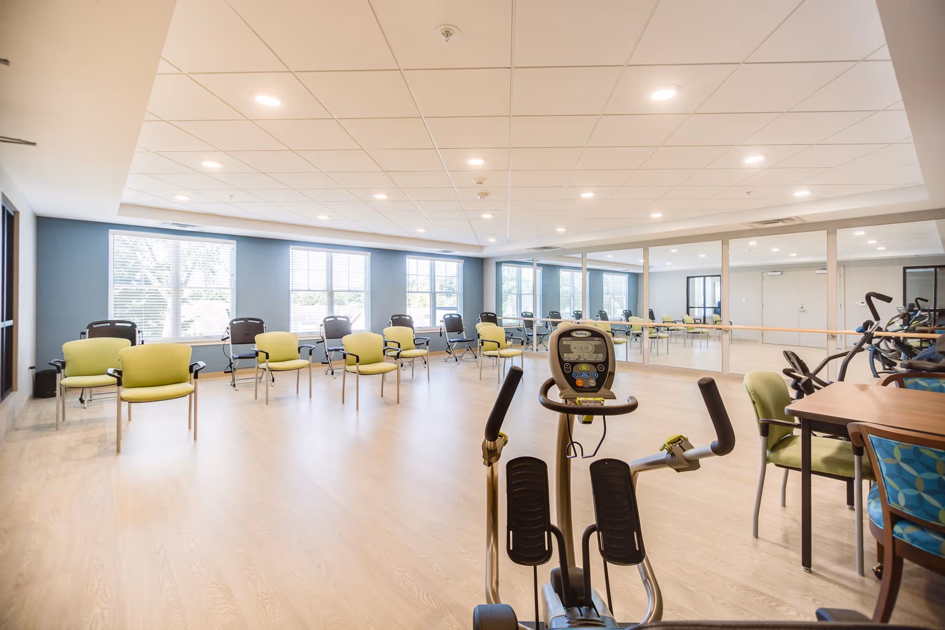 A bright and spacious exercise room with light wood flooring, multiple green and black chairs arranged along the walls, large windows letting in natural light, a mirrored wall with a ballet barre, and exercise equipment including a NuStep machine in the foreground.