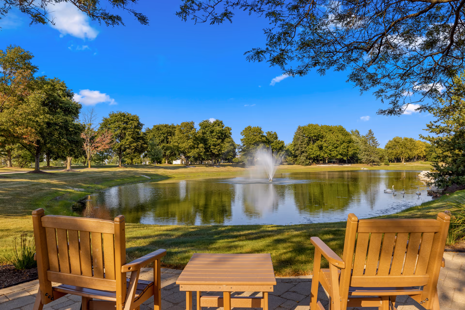 Two wooden chairs and a small wooden table on a paved patio overlooking a pond with a water fountain in the center, surrounded by green grass and trees under a clear blue sky.