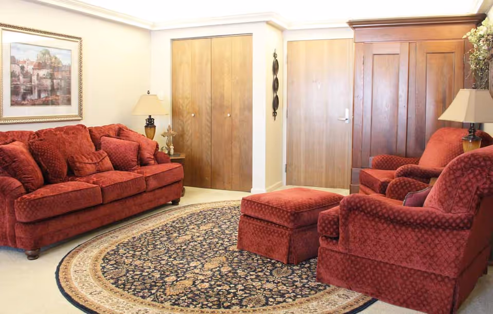 A cozy living room with red upholstered furniture including a sofa, armchair, and ottoman arranged around a round patterned rug. There are two wooden doors and a wooden wardrobe in the background, along with two table lamps and a framed painting on the wall.