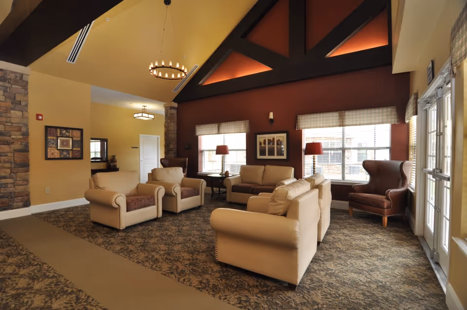 Spacious common room with beige sofas and armchairs, a high vaulted ceiling, and large windows letting in natural light.