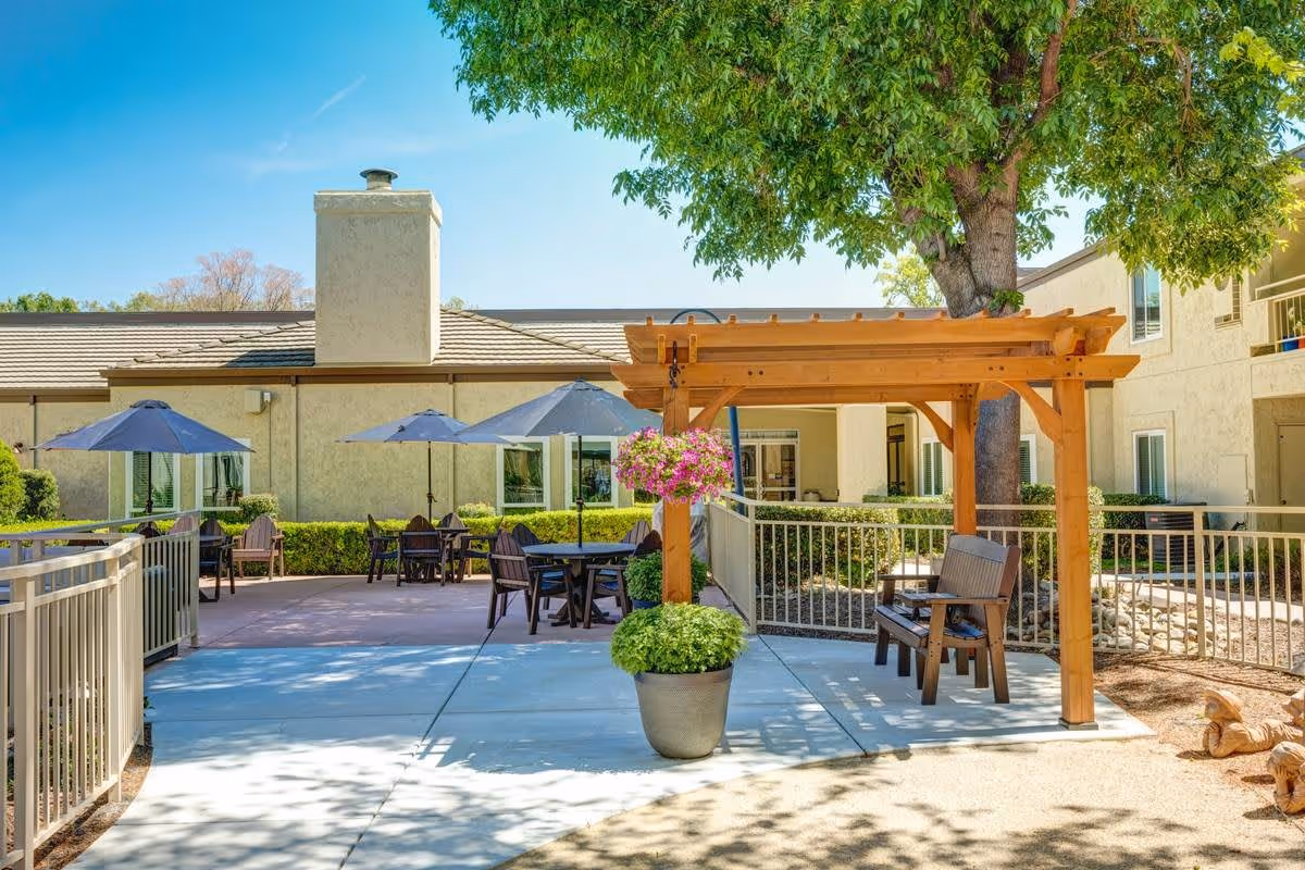 Outdoor patio area at The Oaks at Inglewood featuring a wooden pergola with a bench underneath, a large potted plant with hanging flowers, several tables with blue umbrellas, and a building with beige walls and windows in the background under a clear blue sky.