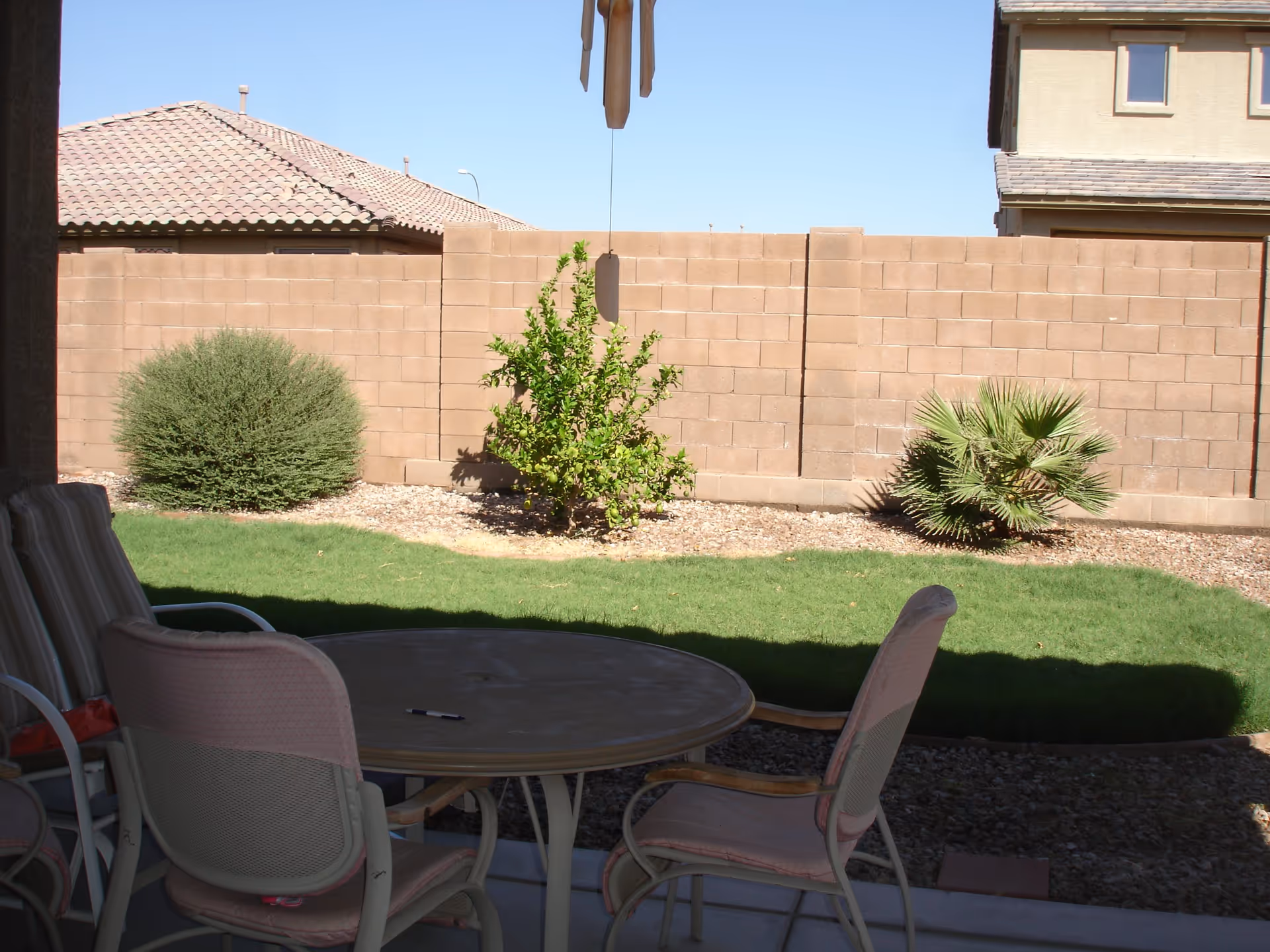 Outdoor patio area with a round table and several chairs with cushions, overlooking a small grassy yard with a few bushes and a brick privacy wall under a clear blue sky.