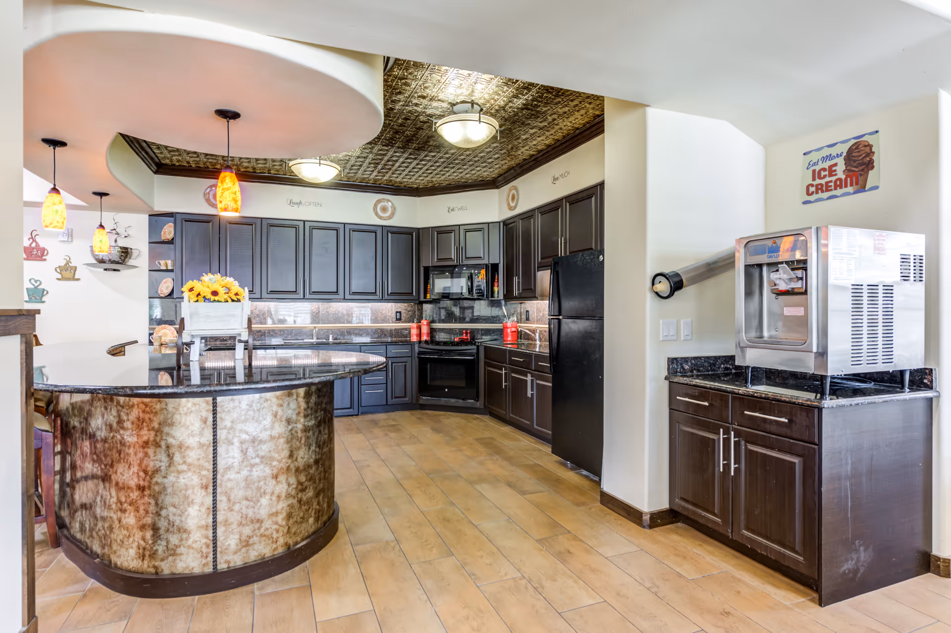 Communal kitchen with a curved island, dark cabinets, pendant lights, and an ice cream machine on a side counter.