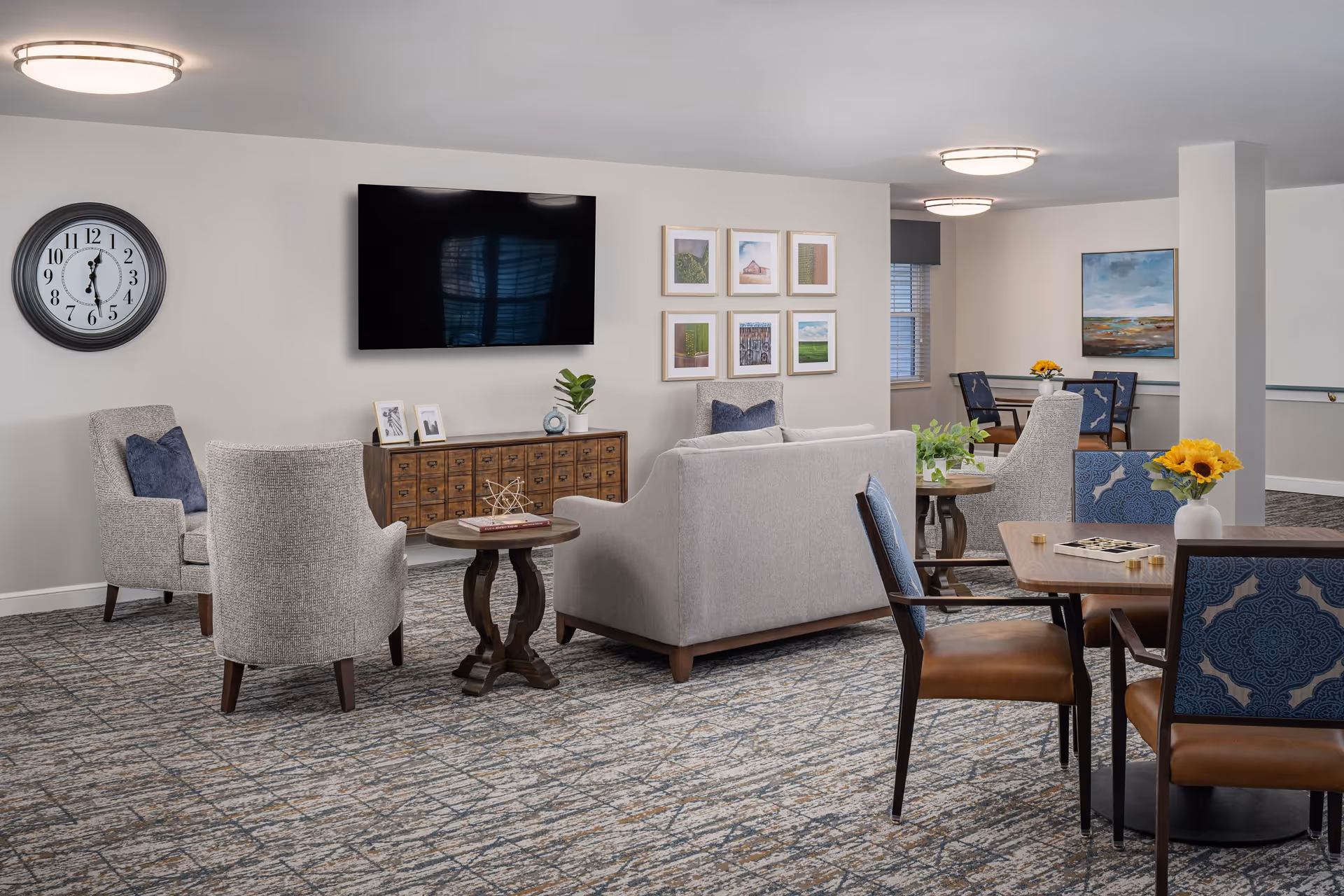 A cozy senior living common area with a gray sofa, two gray armchairs, a wooden side table, and a wall-mounted flat-screen TV. The room features a large wall clock, framed pictures, and a patterned carpet. There are also dining tables with blue and brown chairs, decorated with vases of sunflowers and greenery.