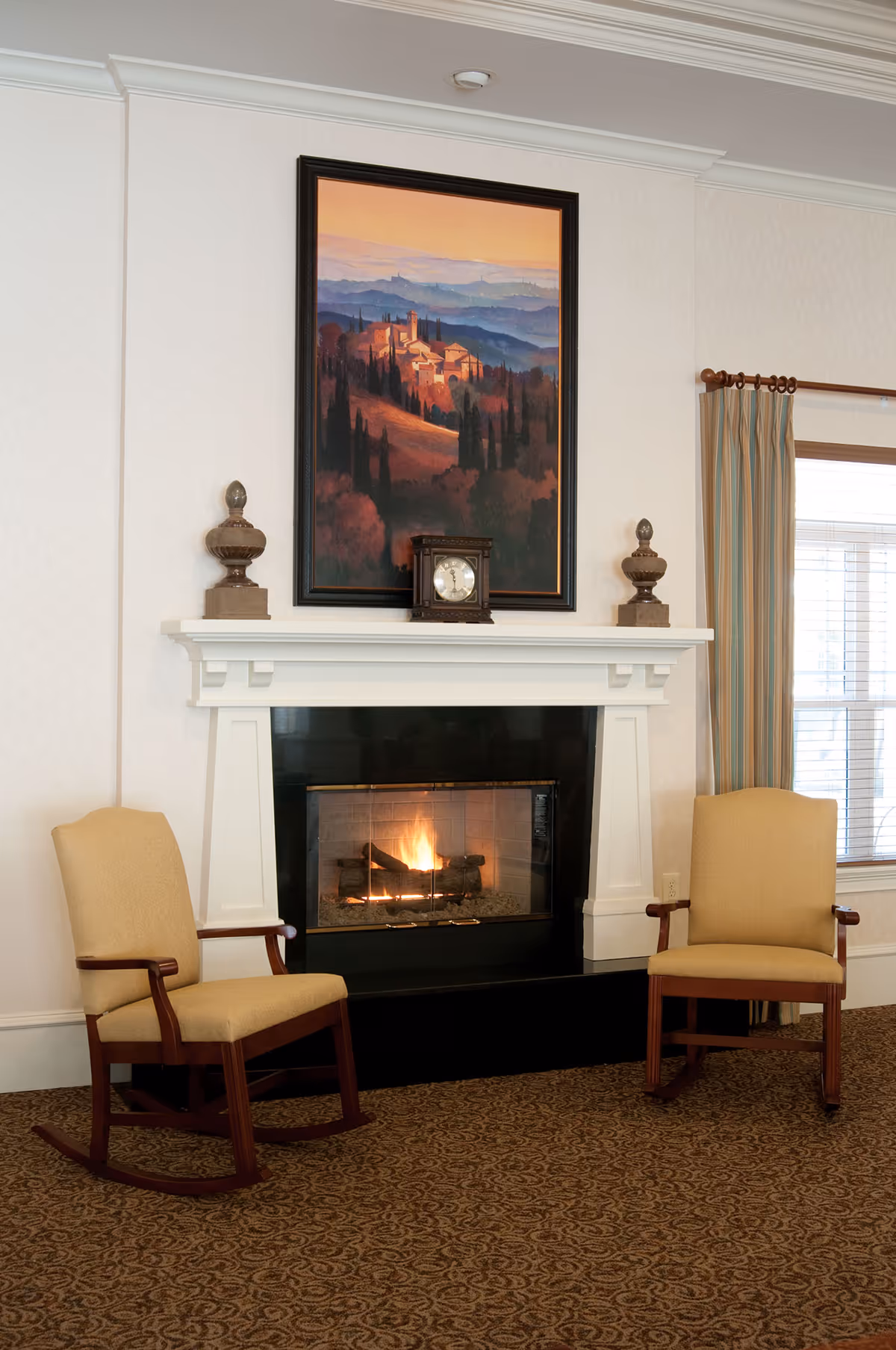 A cozy interior room featuring a lit fireplace with a white mantel. On the mantel, there are two decorative urns and a small clock. Above the mantel hangs a framed painting of a scenic landscape with hills and buildings. Two beige upholstered wooden rocking chairs are positioned on either side of the fireplace. A window with striped curtains is visible on the right side of the image.