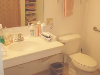 A small bathroom with a white sink and countertop holding various toiletries, a toilet next to the sink, a towel hanging on the wall, and a small trash bin on the floor.