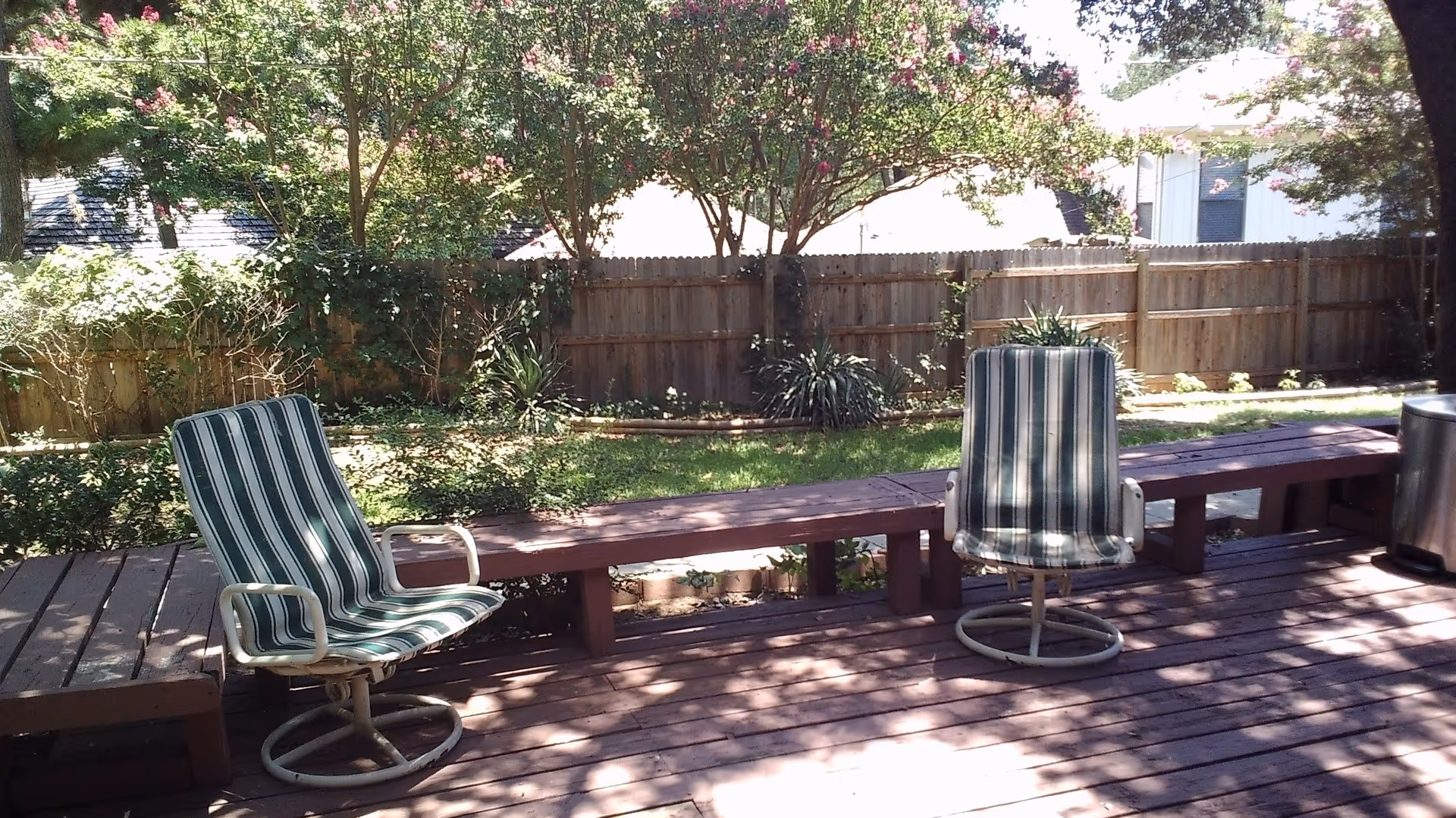 Wooden backyard deck with two striped swivel patio chairs and a long bench in front of a fenced garden.
